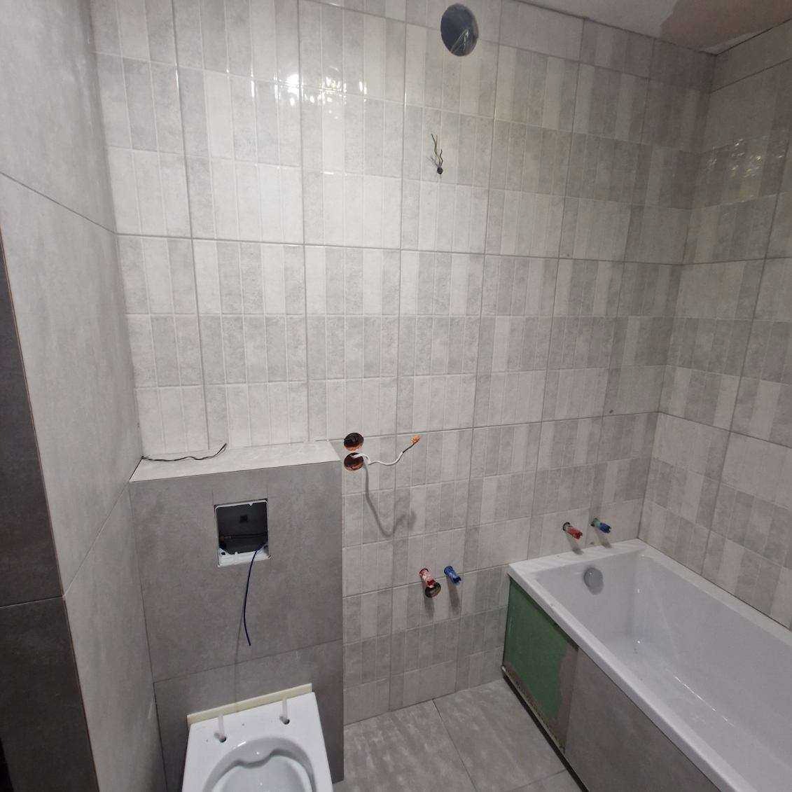 Wykończona łazienka w szarościach z wanną, toaletą i płytkami imitującymi cegłę. Widoczne przyłącza wodne i elektryczne. Minimalistyczny styl.