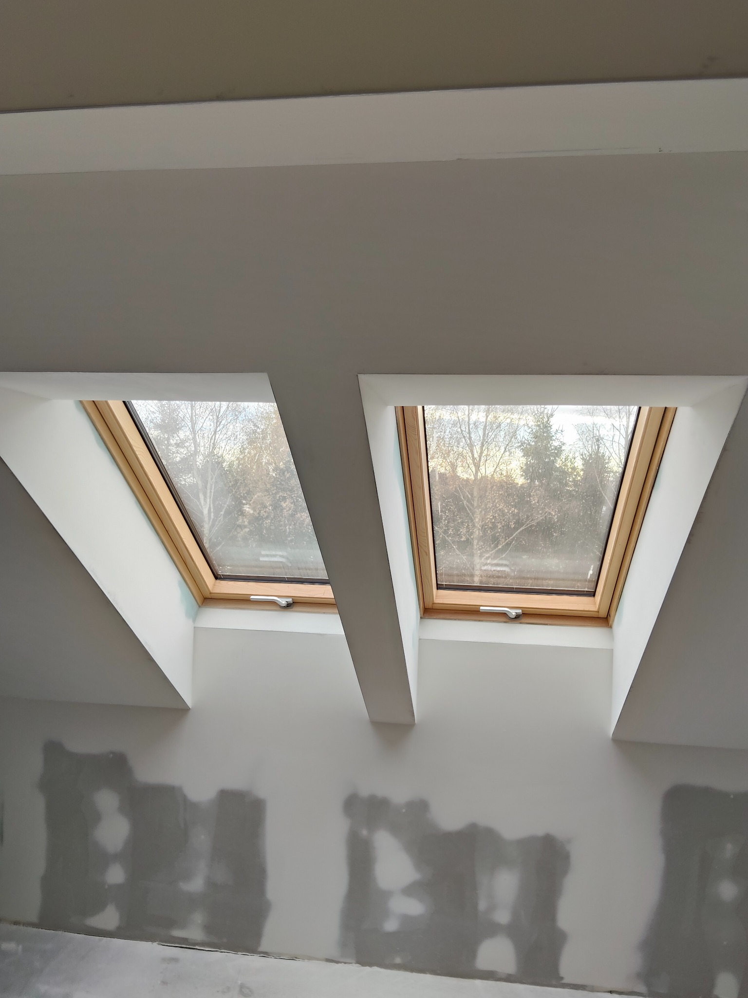 Dwa okna dachowe z drewnianą ramą w ścianie poddasza, widoczne drzewa za oknem. Ściana przygotowana do malowania, z widocznymi śladami szpachlowania.