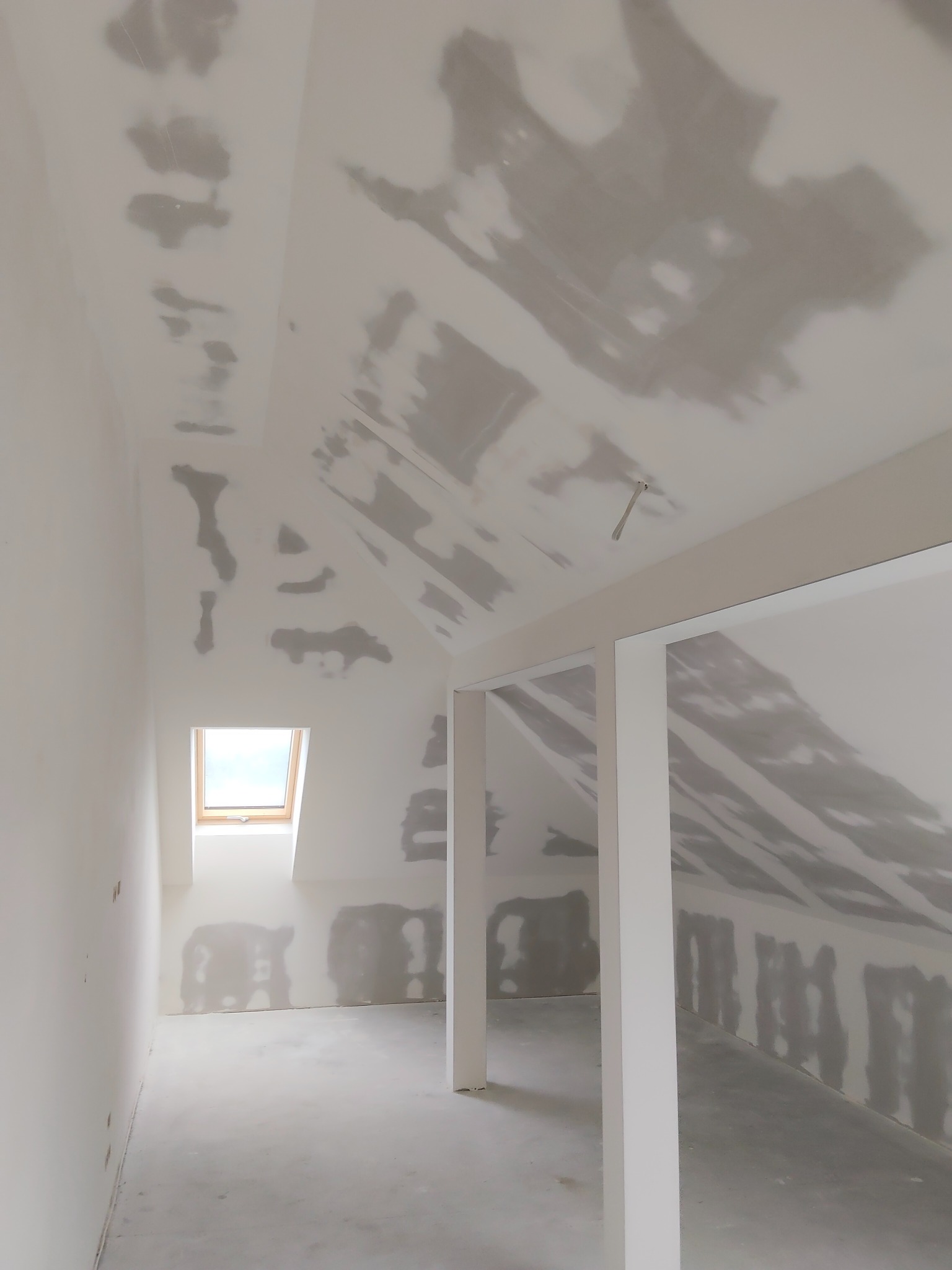 Poddasze ze skosami w trakcie prac wykończeniowych. Widoczne ślady po gładzi na ścianach i suficie. Małe okno dachowe wpuszcza światło.