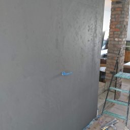 Przemysław Kamiński - Ściana w odcieniach szarości z widoczną fakturą, zabezpieczona taśmą malarską, z fragmentem instalacji elektrycznej i rozstawioną drabiną w tle, na podłodze widoczne ślady farby.