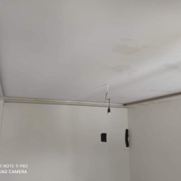 Przemysław Kamiński - Białe ściany i sufit w pokoju w trakcie prac wykończeniowych, widoczne ślady po szpachlowaniu i wystający kabel z żarówką.