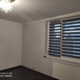 Przemysław Kamiński - Pomalowane na szaro wnętrze pokoju z ciemną podłogą, grzejnikiem i oknem z żaluzjami dzień-noc oraz kryształowym żyrandolem.