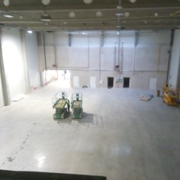 Przemysław Kamiński - Przestronne wnętrze hali produkcyjnej z betonową posadzką, dwoma podnośnikami nożycowymi i pracownikiem w odblaskowej kamizelce, przygotowane do prac wykończeniowych.