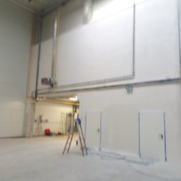 Przemysław Kamiński - Wnętrze hali przemysłowej w trakcie przygotowań do malowania, widoczne zabezpieczone drzwi taśmą malarską, drabina i widoczne instalacje pod sufitem.