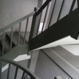 Przemysław Kamiński - Widok z góry na schody z szarą wykładziną i pomalowanymi na szaro poręczami.