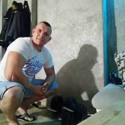 Przemysław Kamiński - Mężczyzna w białej koszulce i klapkach siedzi na kucaka obok ciężarów w pomieszczeniu z szarą ścianą i niebieskim oświetleniem LED.