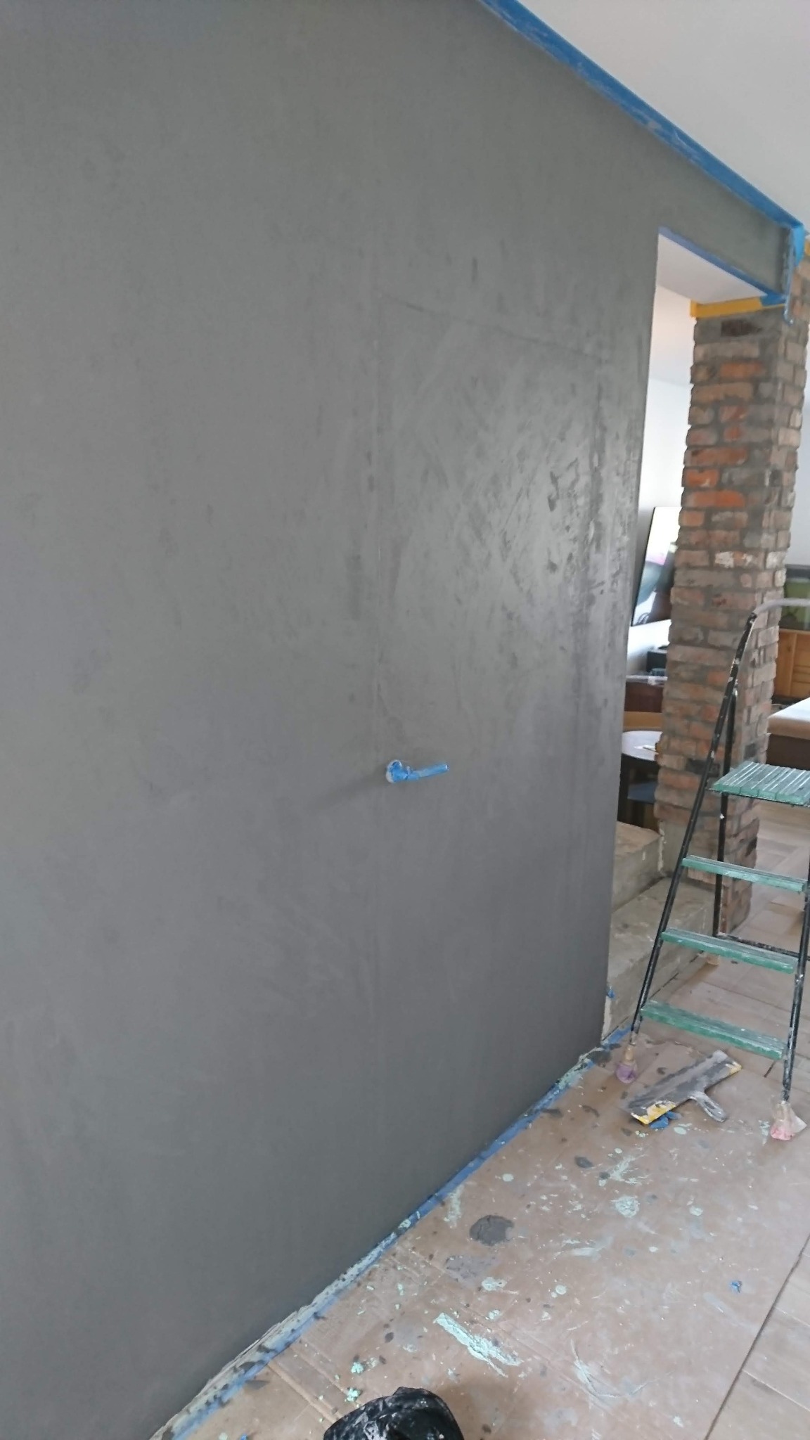 Ściana w odcieniach szarości z widoczną fakturą, zabezpieczona taśmą malarską, z fragmentem instalacji elektrycznej i rozstawioną drabiną w tle, na podłodze widoczne ślady farby.