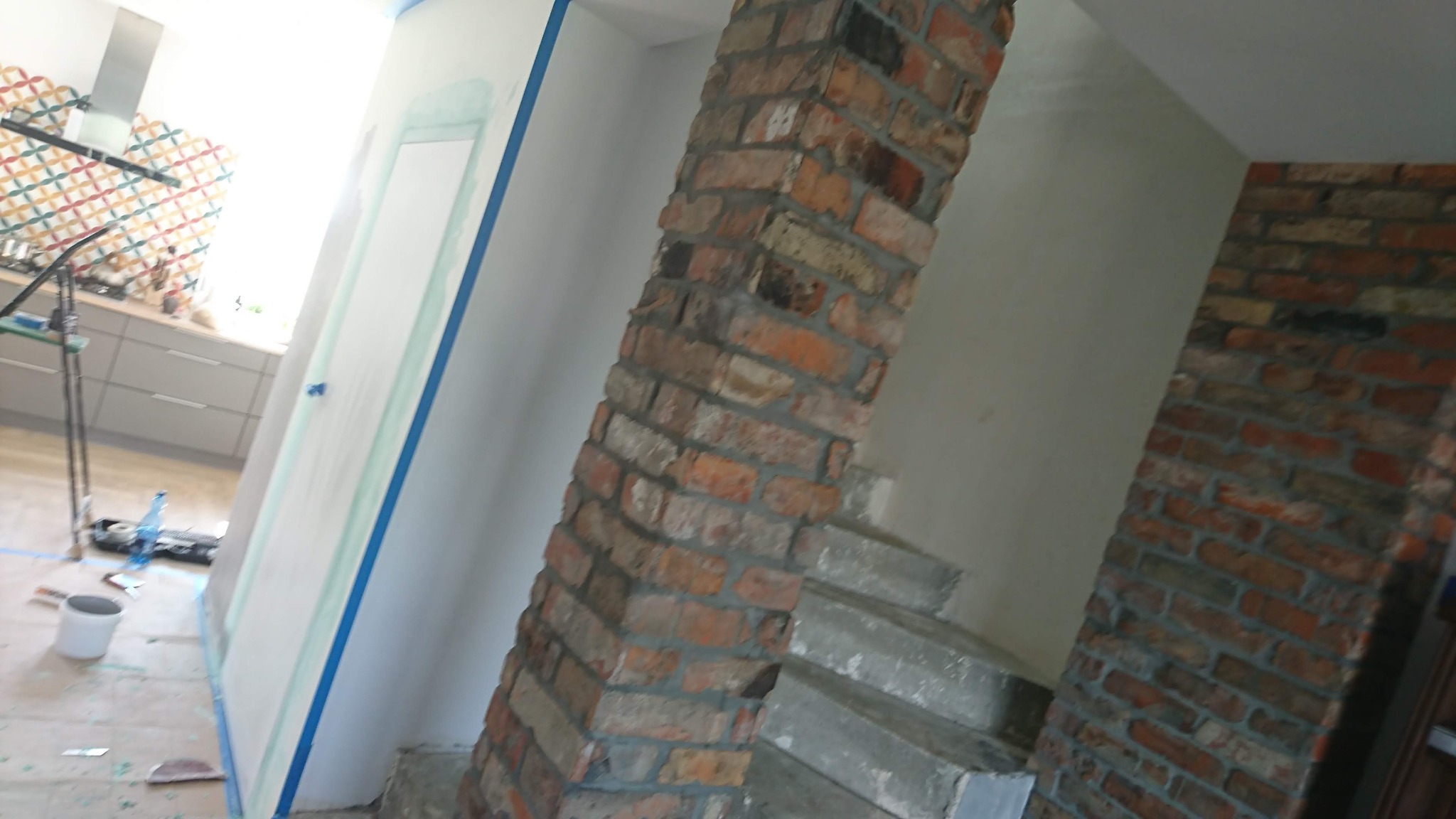 Ściana zabezpieczona niebieską taśmą malarską przed malowaniem, widoczny fragment ceglanej kolumny i schodów w tle kuchnia z kolorową mozaiką.