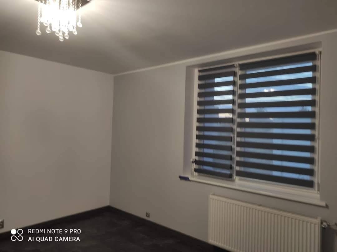 Pomalowane na szaro wnętrze pokoju z ciemną podłogą, grzejnikiem i oknem z żaluzjami dzień-noc oraz kryształowym żyrandolem.