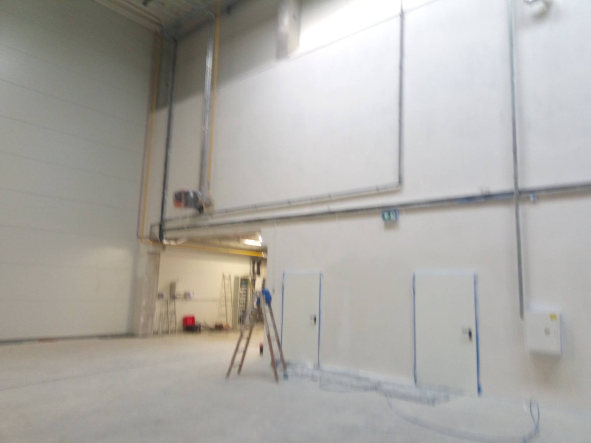 Wnętrze hali przemysłowej w trakcie przygotowań do malowania, widoczne zabezpieczone drzwi taśmą malarską, drabina i widoczne instalacje pod sufitem.