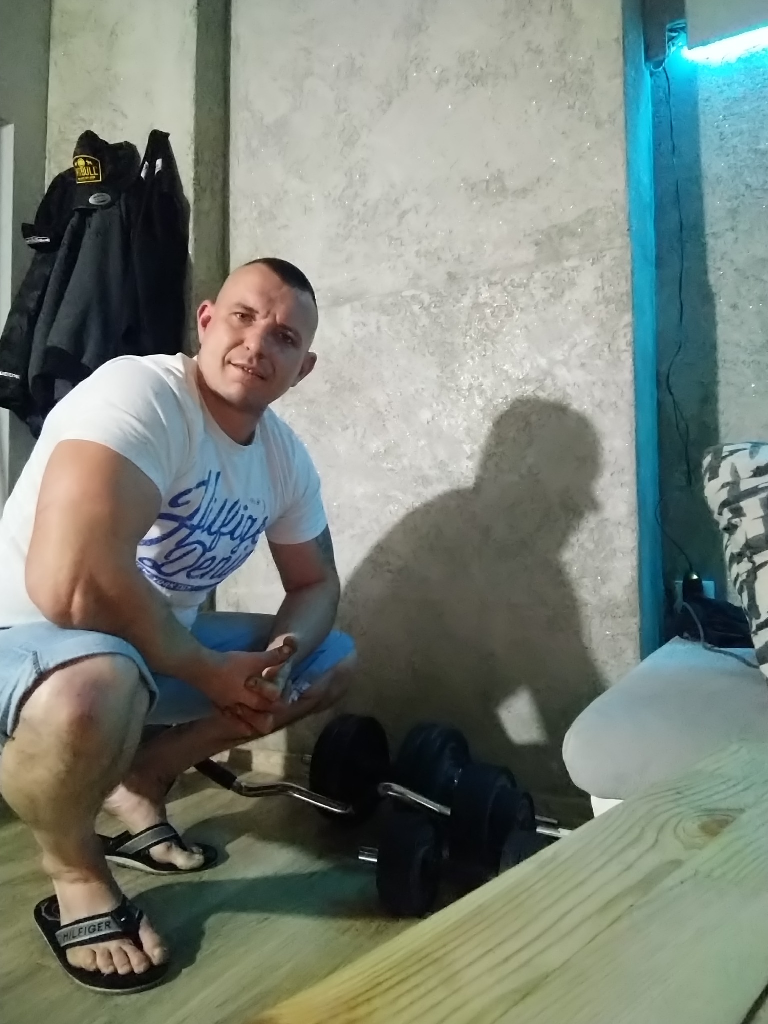 Mężczyzna w białej koszulce i klapkach siedzi na kucaka obok ciężarów w pomieszczeniu z szarą ścianą i niebieskim oświetleniem LED.