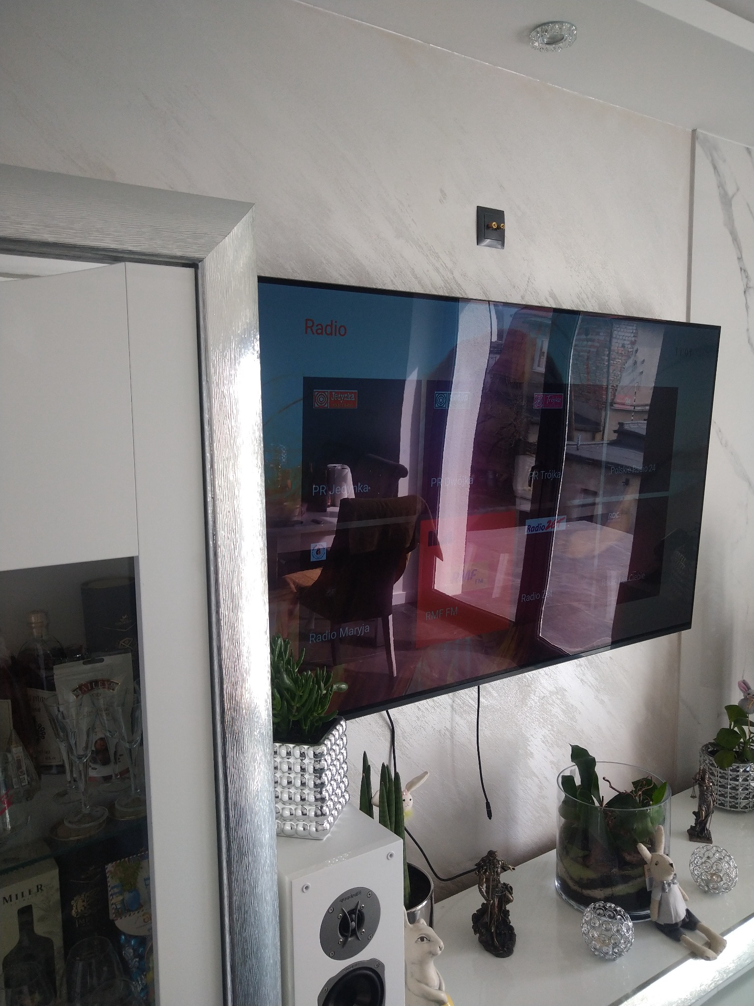 Telewizor LCD zawieszony na ścianie o fakturze imitującej marmur, odbijający w ekranie wnętrze pokoju. Na szafce RTV dekoracje, rośliny w szklanych i metalowych doniczkach oraz głośnik. Widoczny...