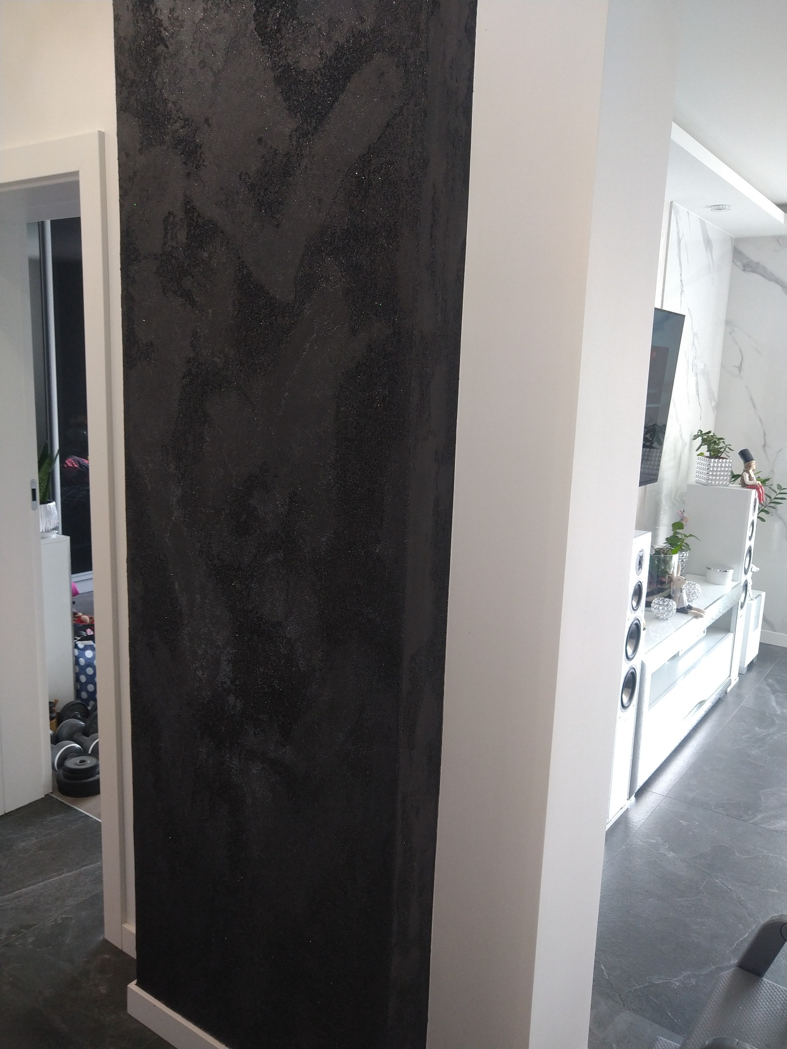 Czarny, teksturowany filar dekoracyjny w nowoczesnym wnętrzu, kontrastujący z białym filarem i listwami przypodłogowymi, widoczny fragment salonu z białymi meblami i marmurową ścianą w tle.