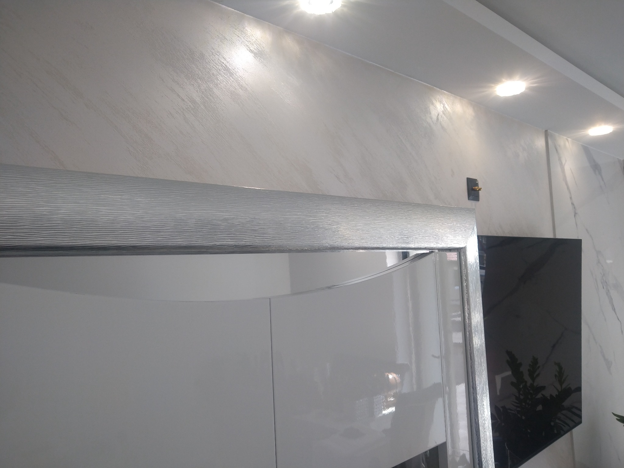 Ściana wykończona dekoracyjną szpachlą w odcieniach beżu i bieli, oświetlona punktowymi lampami sufitowymi, z widocznym fragmentem białej szafy z ozdobną ramą.