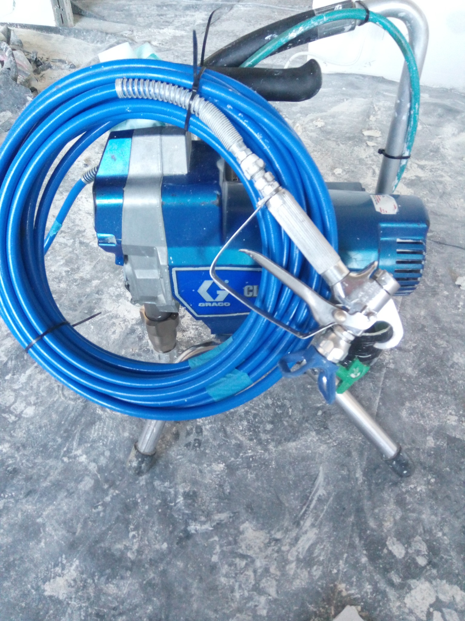 Profesjonalny agregat malarski Graco z niebieskim wężem na szorstkiej, betonowej podłodze pokrytej resztkami farby.