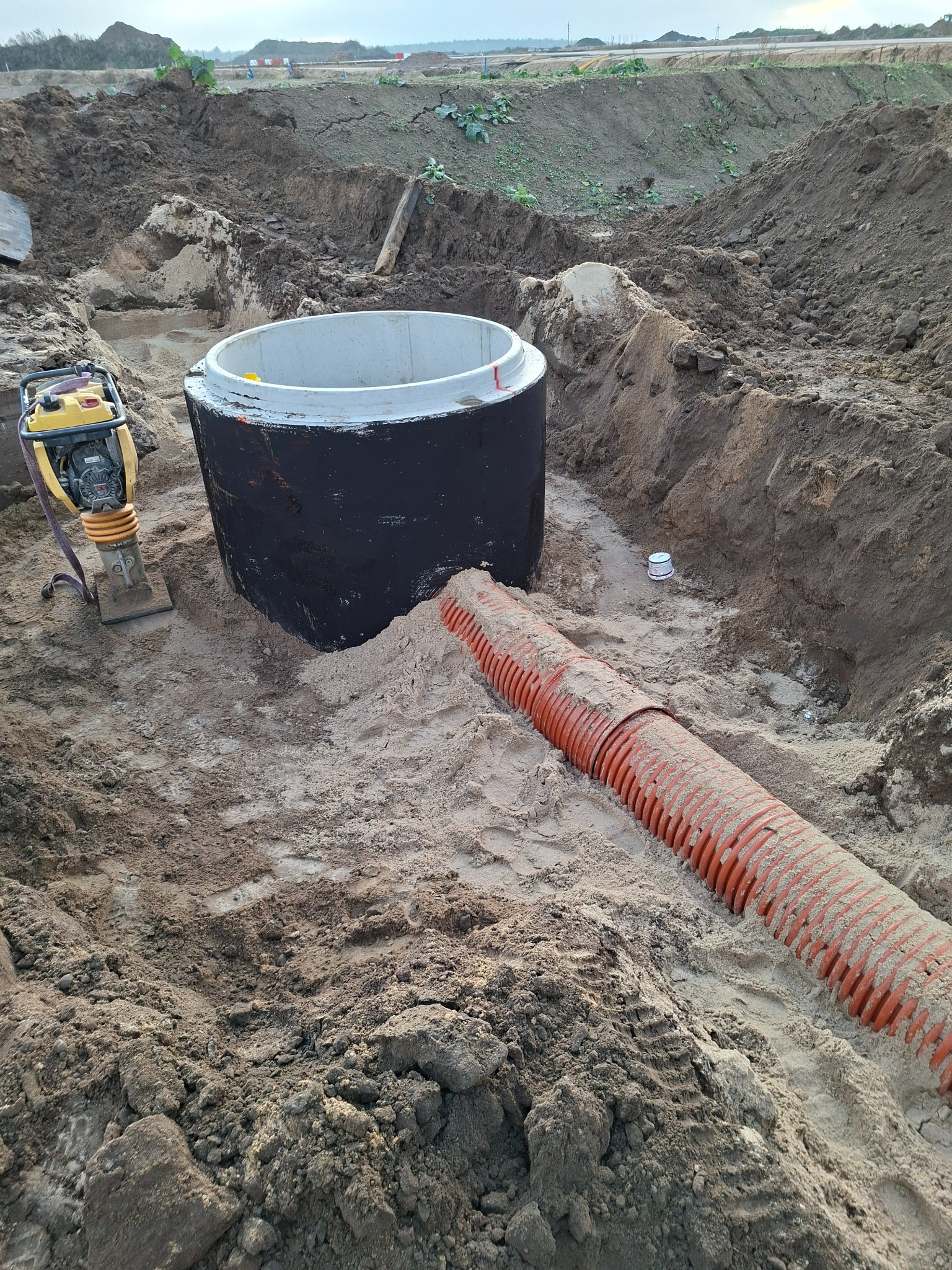 Betonowy zbiornik z podłączoną rurą drenażową w wykopie, obok ubijak wibracyjny. Widoczne ślady prac ziemnych i przygotowanie terenu pod instalację.