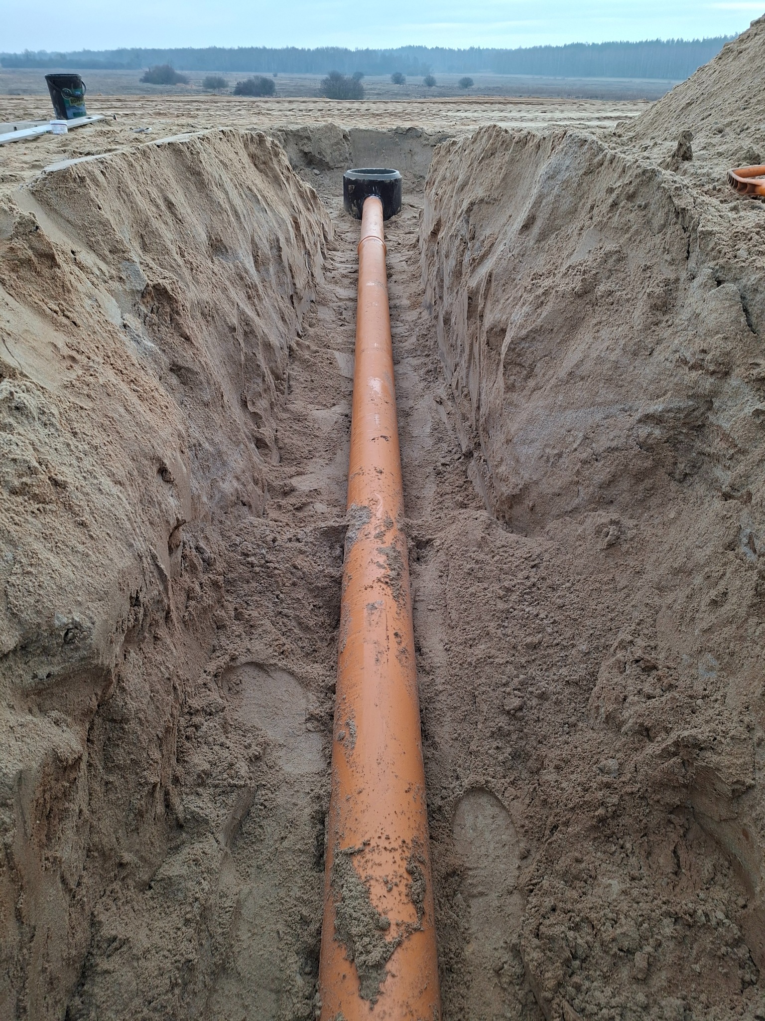 Pomarańczowa rura kanalizacyjna ułożona w wykopie z piasku, widok z góry. Na końcu rury czarna studzienka. W tle pole i las.