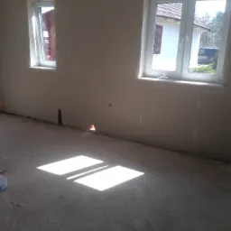 Surowe wnętrze z dwoma oknami, ściany po wstępnym wykończeniu, na podłodze widoczne plamy i fragment worka, wpadające przez okna promienie słoneczne.