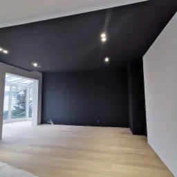 Wnętrze pokoju po malowaniu z czarnym sufitem, jasnymi ścianami i drewnianą podłogą, widoczne instalacje elektryczne na podłodze, okno balkonowe z widokiem na drzewa.