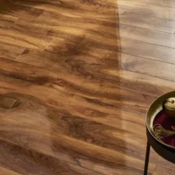 Ułożona podłoga z paneli imitujących ciemne drewno z wyraźnym usłojeniem, częściowo oświetlona, z widocznym fragmentem czarnego stolika z dekoracyjną misą i ozdobami.