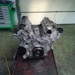 Zdemontowany silnik samochodowy V-kształtny, widoczne głowice, wałki rozrządu z kołami zębatymi i łańcuchem, umieszczony na metalowym wózku warsztatowym, w tle zielony wózek narzędziowy TOPTUL...