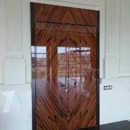 Dwuskrzydłowe drzwi z egzotycznego drewna o geometrycznym wzorze, z widocznym odbiciem otoczenia i nowoczesną klamką, osadzone w klasycznej, białej ramie z pilastrami.