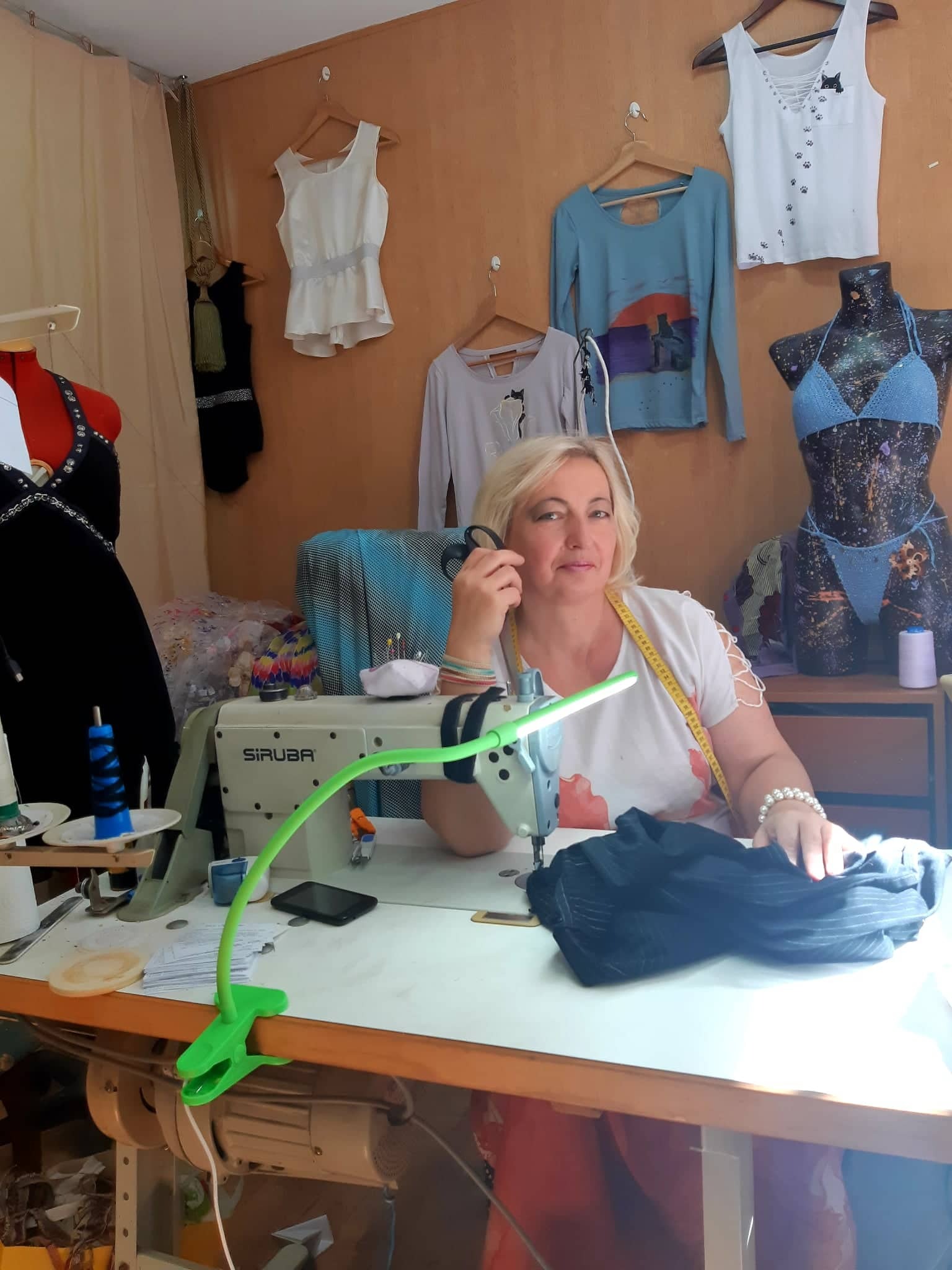 Krawcowa z blond włosami siedzi przy maszynie do szycia Siruba, w tle wieszaki z damską odzieżą i manekin w błękitnym stroju kąpielowym, na stole czarny materiał w paski.