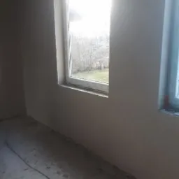 Świeżo otynkowane ściany wewnętrzne w pomieszczeniu z dwoma oknami, widoczne niedoskonałości na podłodze przed dalszym wykończeniem.