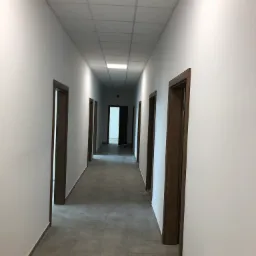 Długi korytarz biurowy z białymi ścianami, szarą podłogą z płytek i drewnianymi ościeżnicami drzwi, widoczny grzejnik zabezpieczony folią.