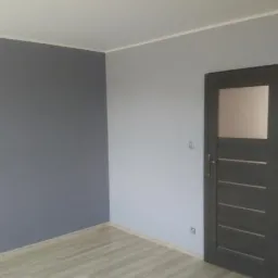 Pomieszczenie po malowaniu ścian na dwa kolory: szary i jasnoszary, z widocznymi panelami podłogowymi i ciemnymi drzwiami z szybą.