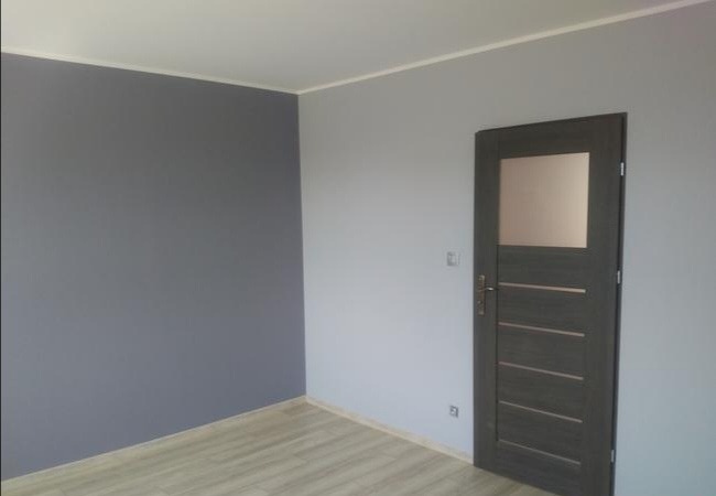 Pomieszczenie po malowaniu ścian na dwa kolory: szary i jasnoszary, z widocznymi panelami podłogowymi i ciemnymi drzwiami z szybą.