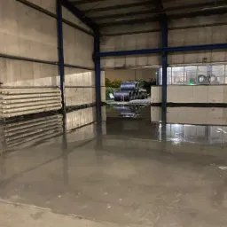 Wnętrze hali z połyskującą, mokrą posadzką i zwojami stali zbrojeniowej w tle.