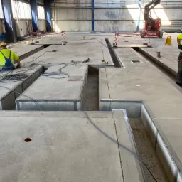 Betonowa wylewka w hali przemysłowej z wyciętymi kanałami, pracownicy w żółtych kamizelkach i kaskach, sprzęt budowlany w tle.