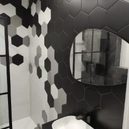 Nowoczesna łazienka z heksagonalnymi płytkami w odcieniach szarości i czerni, okrągłe lustro odbija fragment kabiny prysznicowej, biała umywalka nablatowa o owalnym kształcie z czarną baterią.
