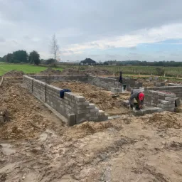 Budowa fundamentów domu z bloczków betonowych: murarz w czerwonej czapce układa bloczki, widoczne zbrojenia i drewniane szalunki, w tle krajobraz z domem i drzewami.