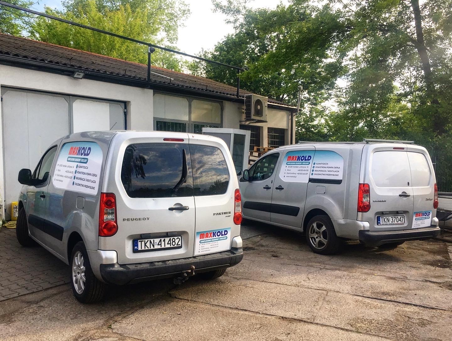 Dwa srebrne samochody dostawcze Peugeot Partner firmy MAXKOLD zaparkowane przed budynkiem, z reklamą usług klimatyzacji, pomp ciepła i rekuperacji na tylnych szybach.