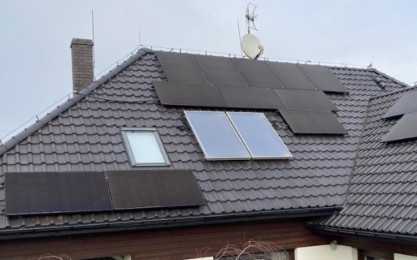 Czarny dach pokryty dachówką z zamontowanymi panelami słonecznymi i kolektorami słonecznymi, antena satelitarna i komin w tle pochmurnego nieba.