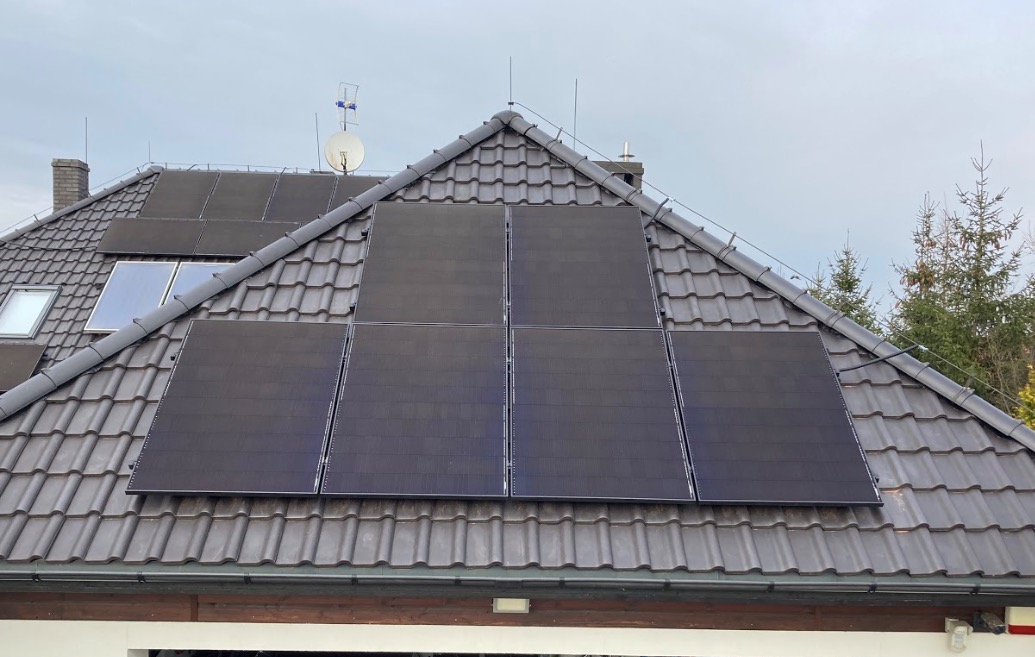 Cztery panele słoneczne w układzie 2x2 na ciemnym, spadzistym dachu w Kryniczno, z dodatkowymi panelami i anteną satelitarną widocznymi w tle, na tle pochmurnego nieba.