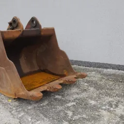 Używana, rdzawa łyżka koparki z widocznymi śladami eksploatacji i hydraulicznym młotem wyburzeniowym obok, na betonowym podłożu przy szarej ścianie budynku.