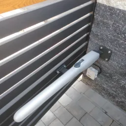 Szczegółowe ujęcie automatycznego napędu do bramy, zamontowanego na nowoczesnej, poziomej bramie z szarych profili, z widoczną kostką brukową i fragmentem kamiennej elewacji.