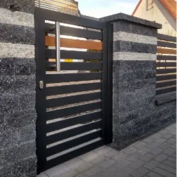 Nowoczesna furtka z poziomymi, czarnymi profilami, wbudowana w ogrodzenie z szarego, łupanego kamienia z poziomymi, białymi pasami, widoczna srebrna klamka i zamek.