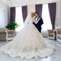 Panna młoda w koronkowej sukni i welonie, oraz pan młody w ciemnym garniturze, stoją blisko siebie w eleganckim wnętrzu z marmurową podłogą i stylizowanymi fotelami.