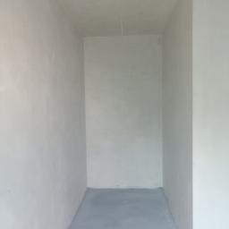 Usługi Tynkarskie Wojciech Kała - Wnętrze małego pomieszczenia po nałożeniu jasnoszarego tynku na ściany i wylaniu szarej posadzki. Widoczny kabel zwisający z sufitu.