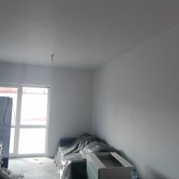 Usługi Tynkarskie Wojciech Kała - Wnętrze po nałożeniu jasnych, gładkich tynków; meble zabezpieczone folią malarską, widoczne gniazdka elektryczne.