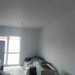 Wnętrze po nałożeniu jasnych, gładkich tynków; meble zabezpieczone folią malarską, widoczne gniazdka elektryczne.