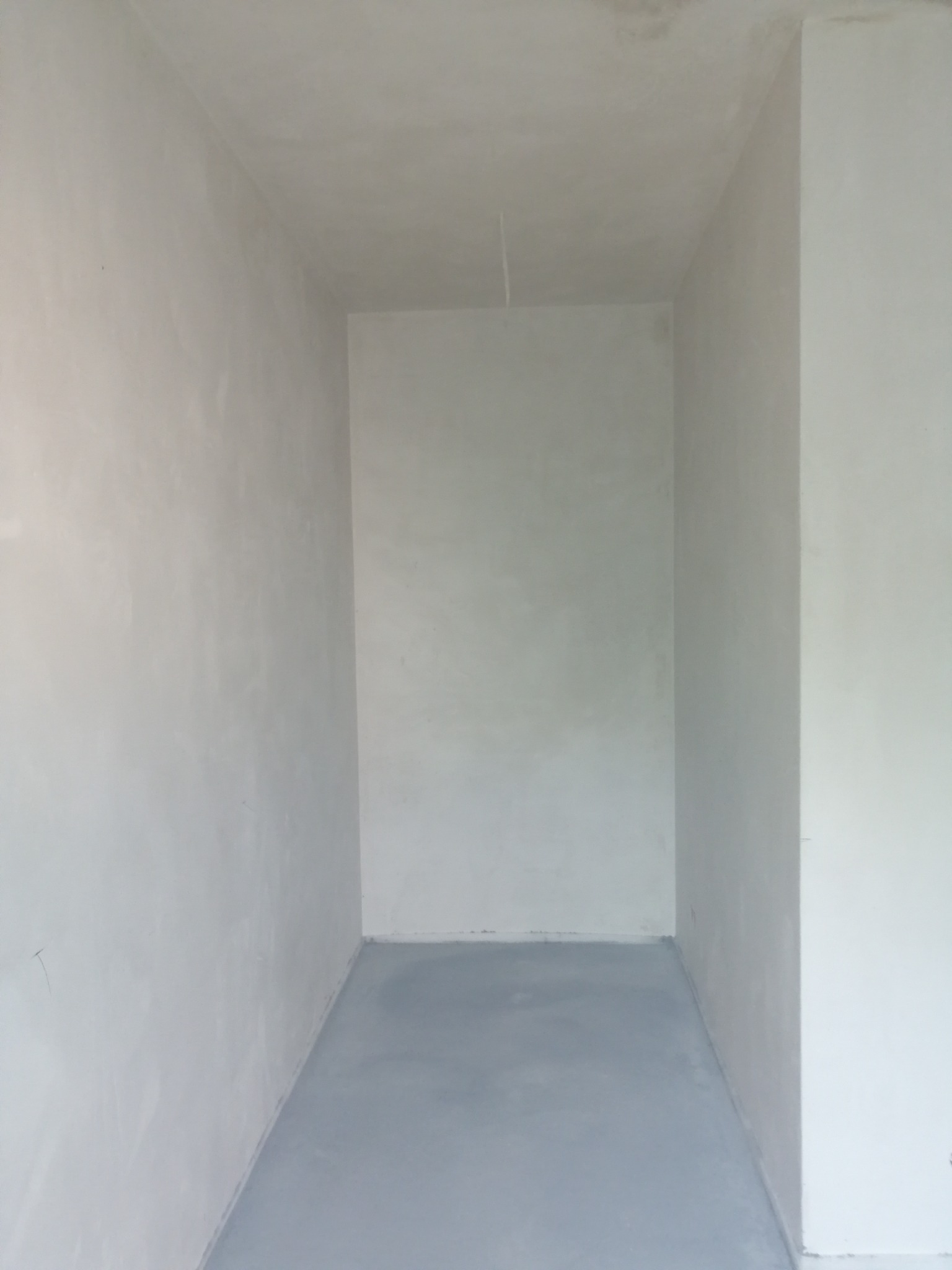 Wnętrze małego pomieszczenia po nałożeniu jasnoszarego tynku na ściany i wylaniu szarej posadzki. Widoczny kabel zwisający z sufitu.