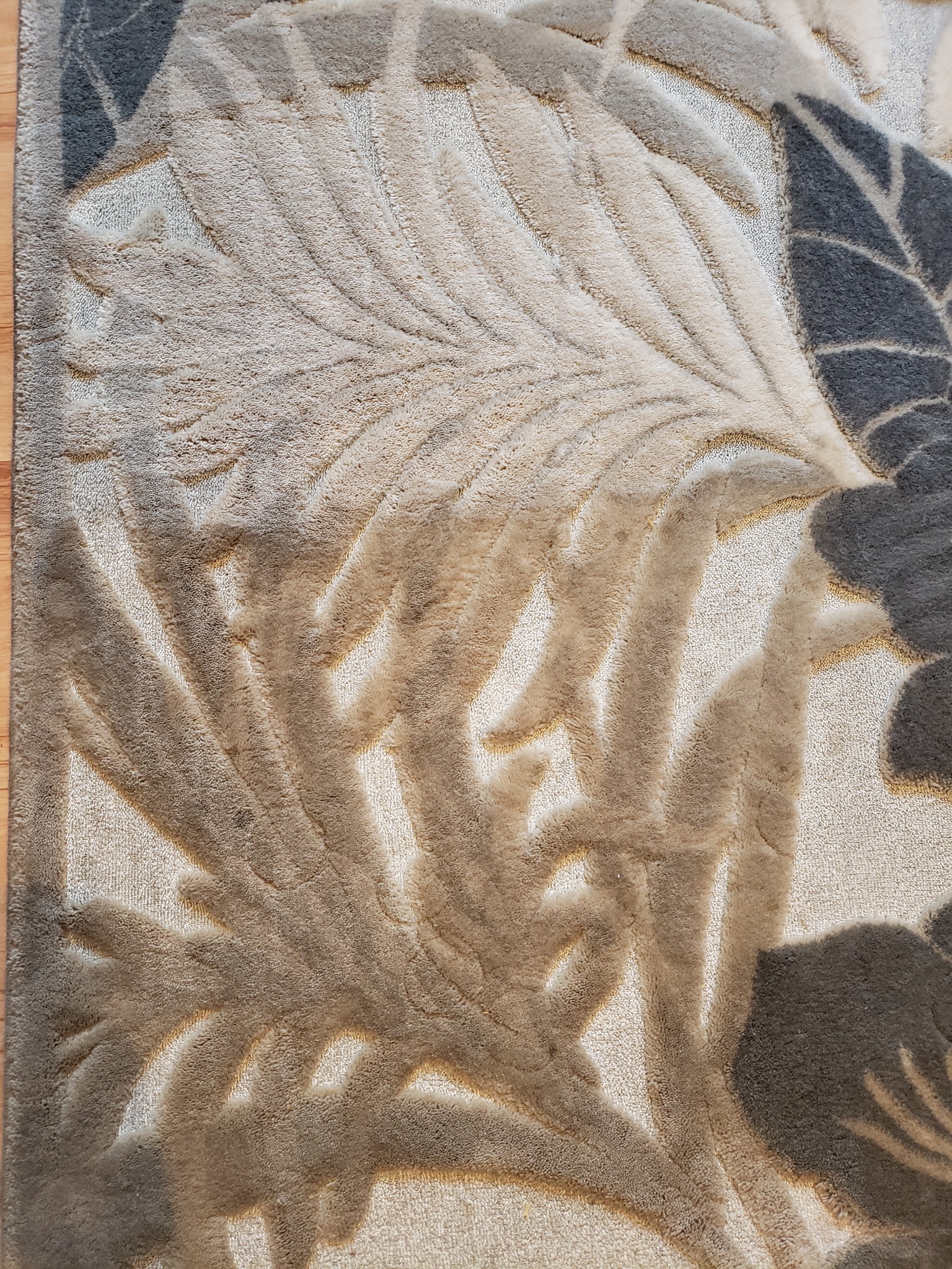 Beżowy dywan z wytłaczanym wzorem liści i kwiatów w odcieniach szarości, leżący na drewnianej podłodze, widziany z góry.