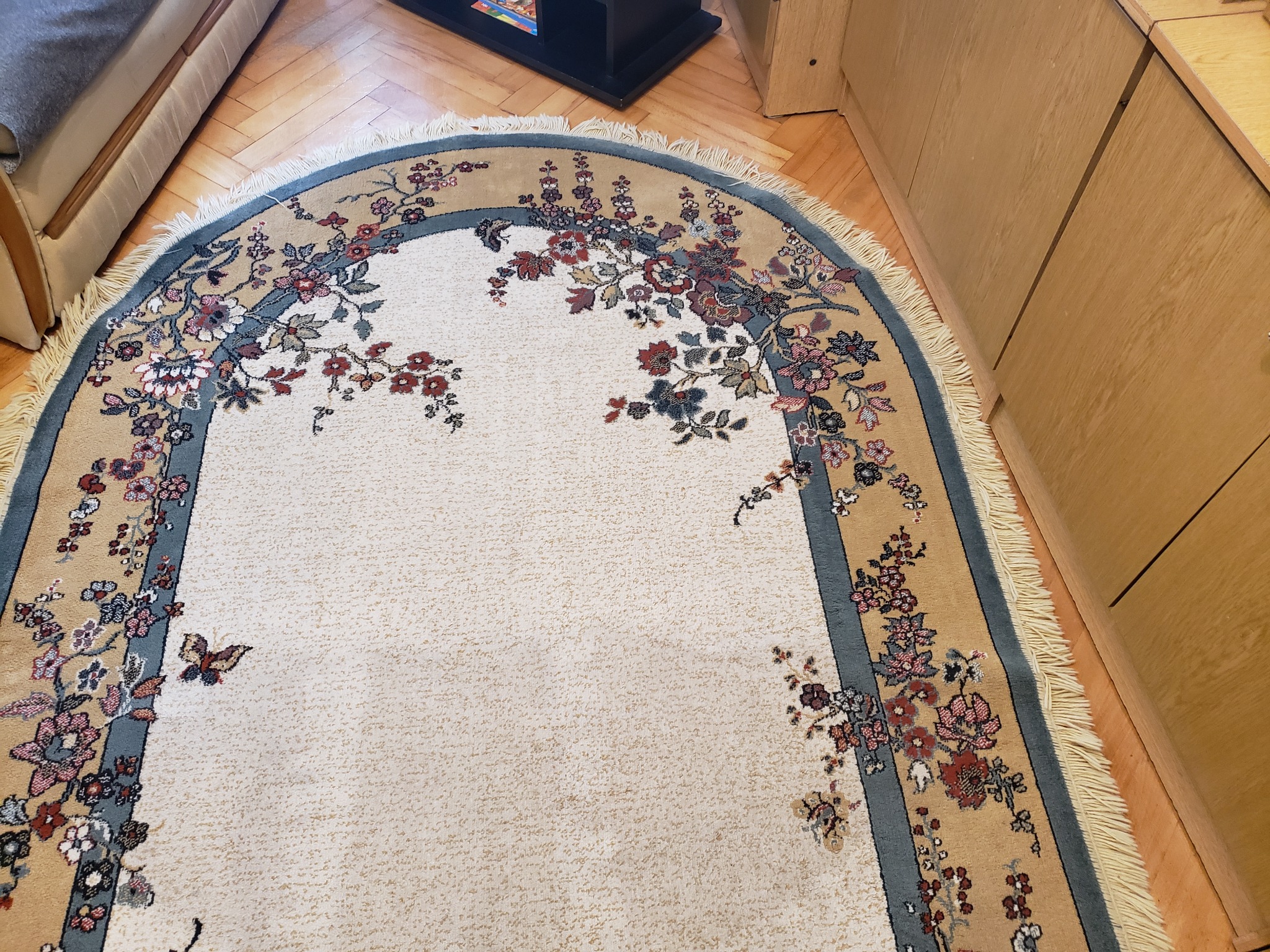 Owalny dywan z kremowym tłem i floralnym wzorem w odcieniach beżu, brązu i szarości, leżący na drewnianej podłodze w jasnym wnętrzu.