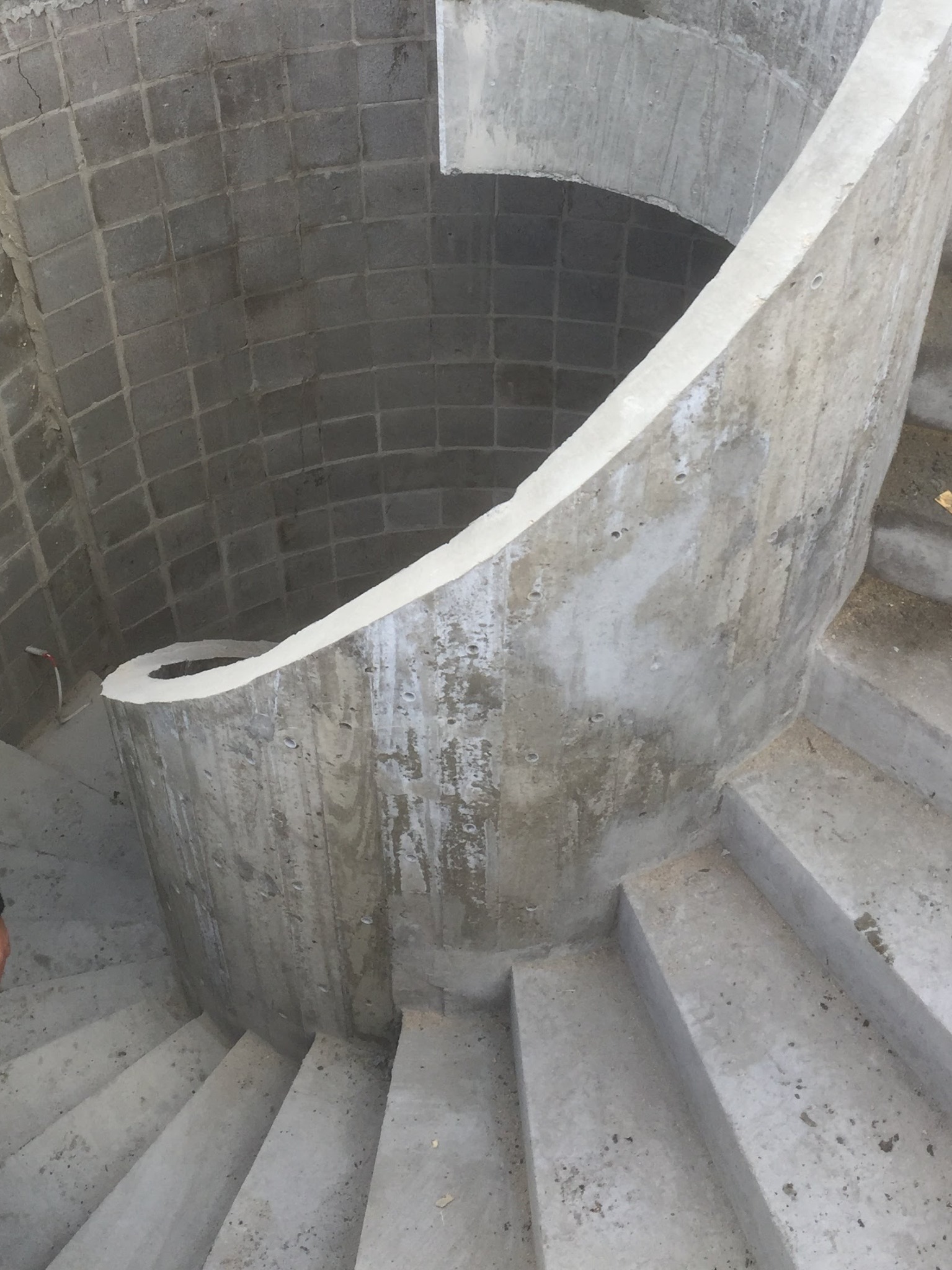 Wewnętrzne spiralne schody w surowym betonie, z widoczną fakturą szalunku i blokami betonowymi w tle, ukazujące etap budowy.