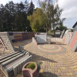 Ekspozycja różnorodnych materiałów nawierzchniowych na zewnątrz, z ułożonym okręgiem z kolorowej kostki w centrum, otoczona stojakami z próbkami i elementami dekoracyjnymi, na tle drzew i budynku.